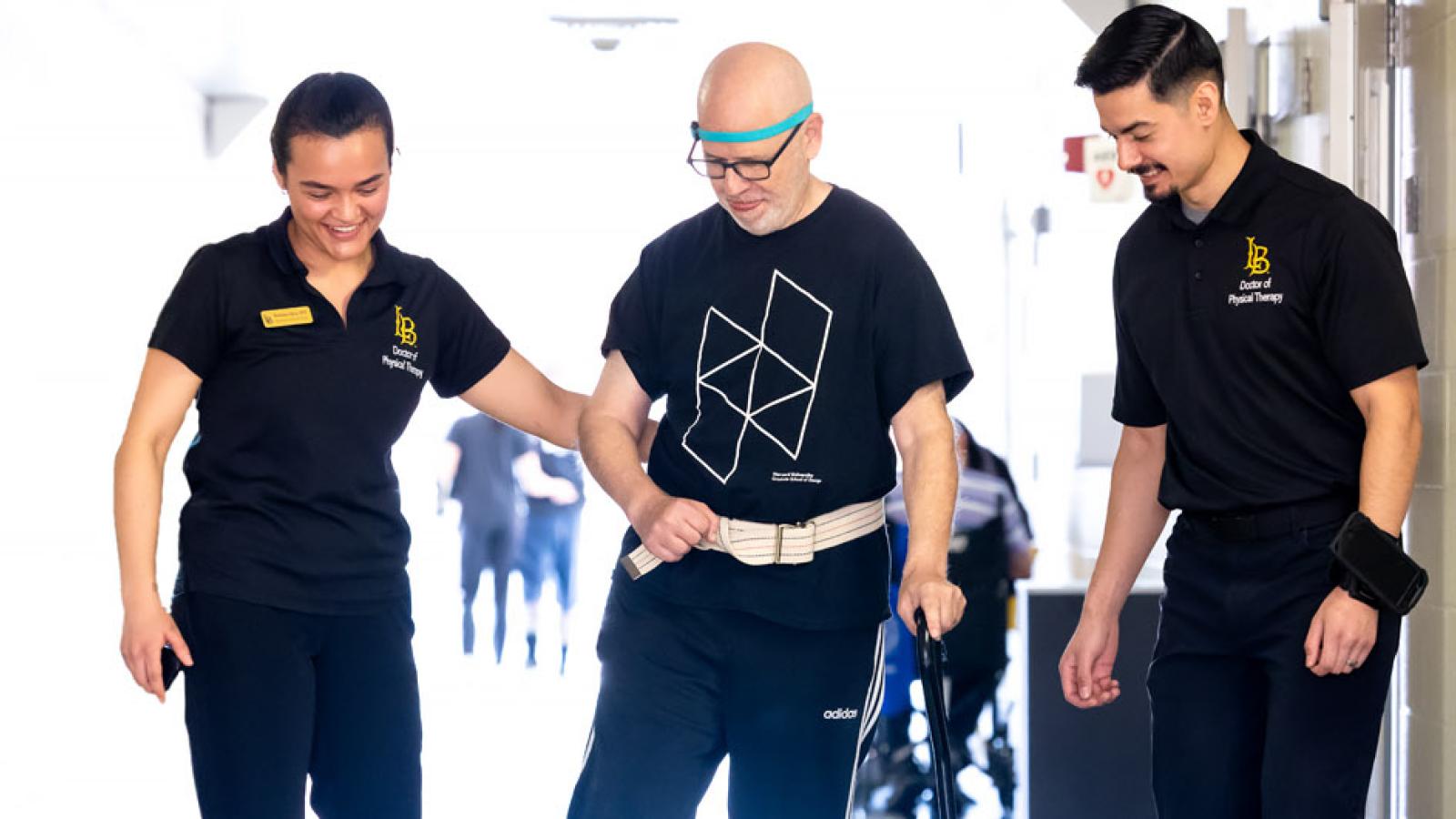 Two CSULB students walk beside a Neuro Lab client as he uses a cane to walk down a hallway.