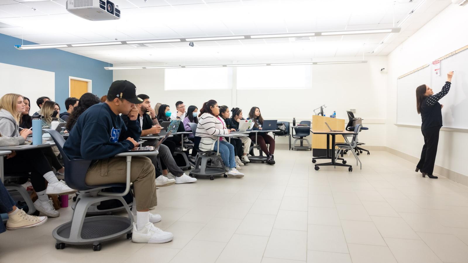 Professor writing on whiteboard with class of students watching.