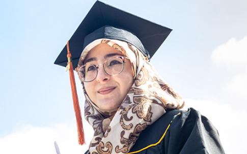 Student wearing hijab and graduation gap holds notepad and water bottle