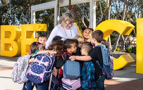 Long Beach State University President Jane Close Conoley stands among a group of children with backpacks who are hugging her in front of large yellow "GO BEACH" letters on campus.