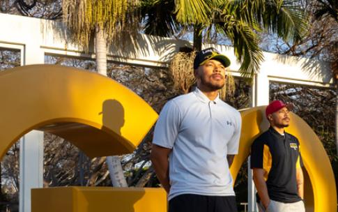 Alumni Markel and Martel Cooper stand in front of large yellow "GO BEACH" letters at Long Beach State 