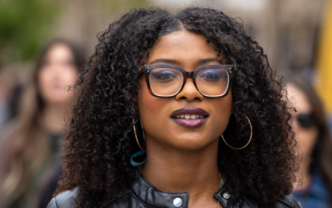 A person with curly hair, glasses and a leather jacket looks ahead while walking outdoors