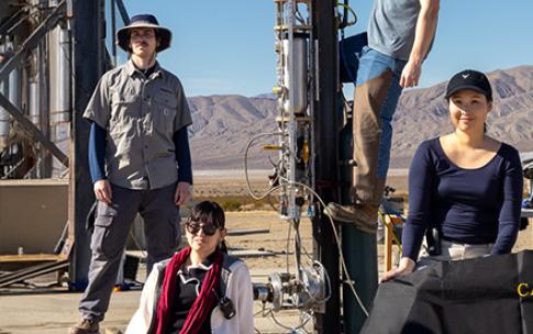 Four people pose next to a vertical test stand with piping and equipment in desert