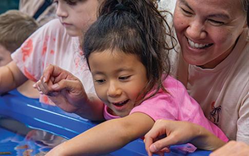 A child touches marine life in a tank while an adult supports them from behind