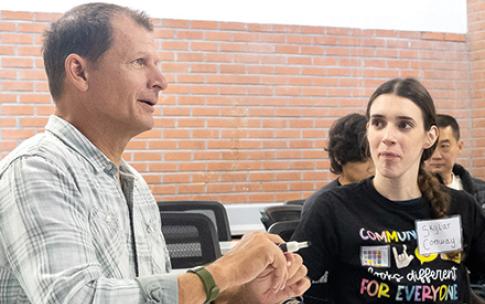 Two people talk at a desk with whiteboards during class