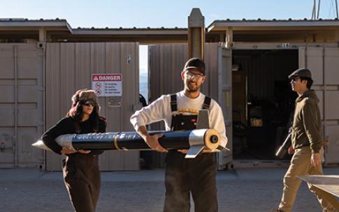 Two students carry a rocket body across a sandy outdoor workspace while others walk nearby.