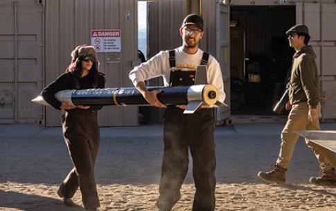 Two students carry a rocket body across a sandy outdoor workspace while others walk nearby.