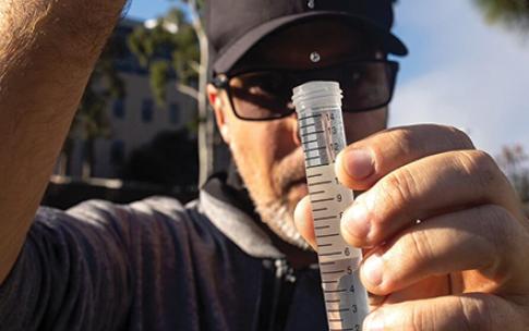 A person holds a syringe over a clear tube, releasing drops of liquid during outdoor sample collection.