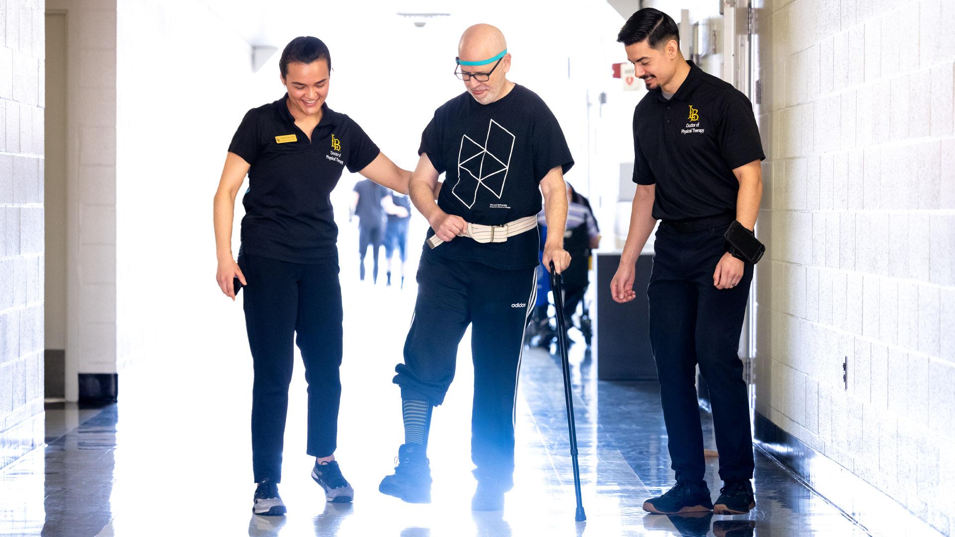 Students wearing black help a Neuro Clinic client walk down a hallway as he uses a cane to walk