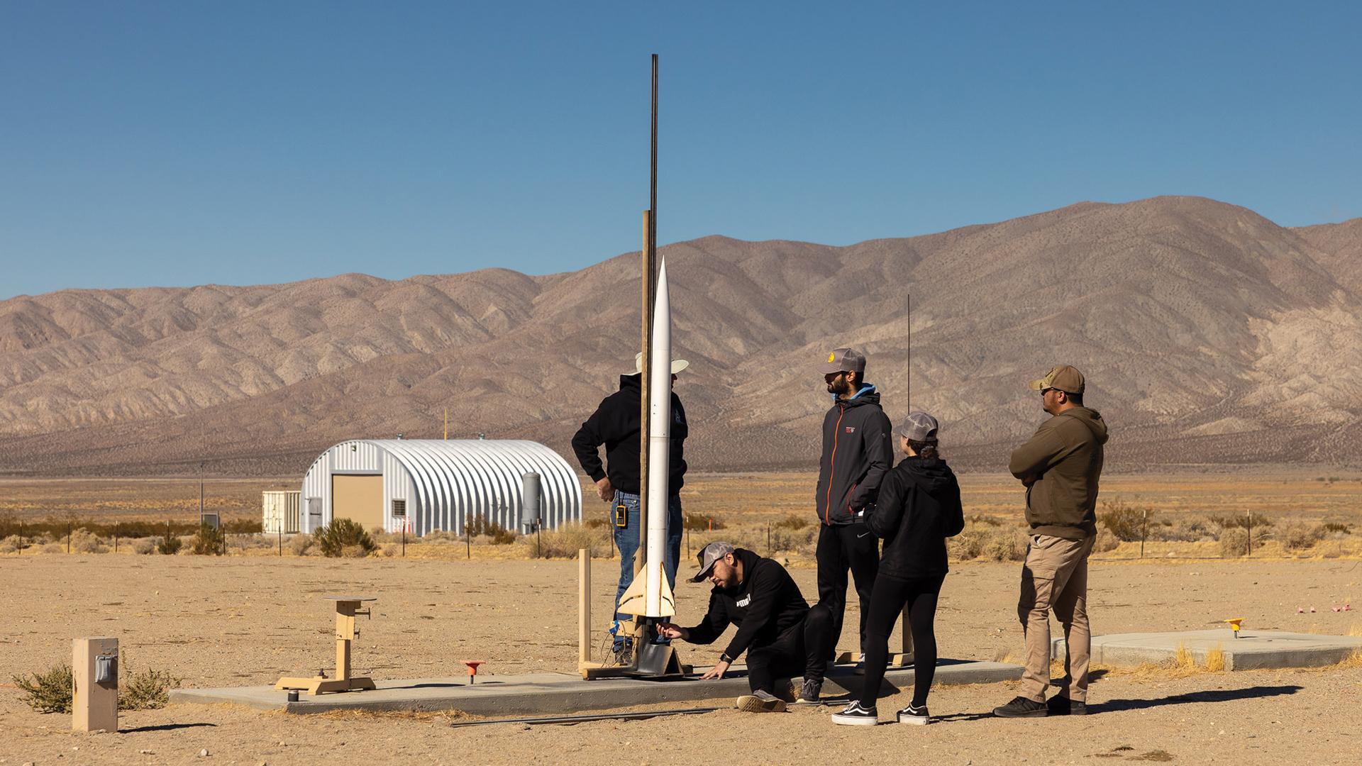Six people stand around a tall model rocket on a desert launch pad near a metal building with mountains in the background