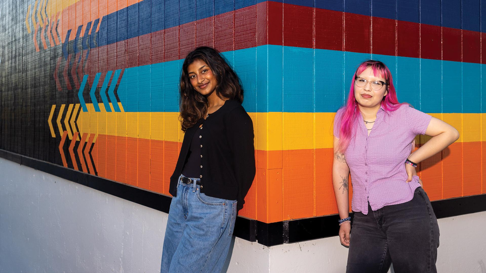 Two students lean against a brightly colored striped wall