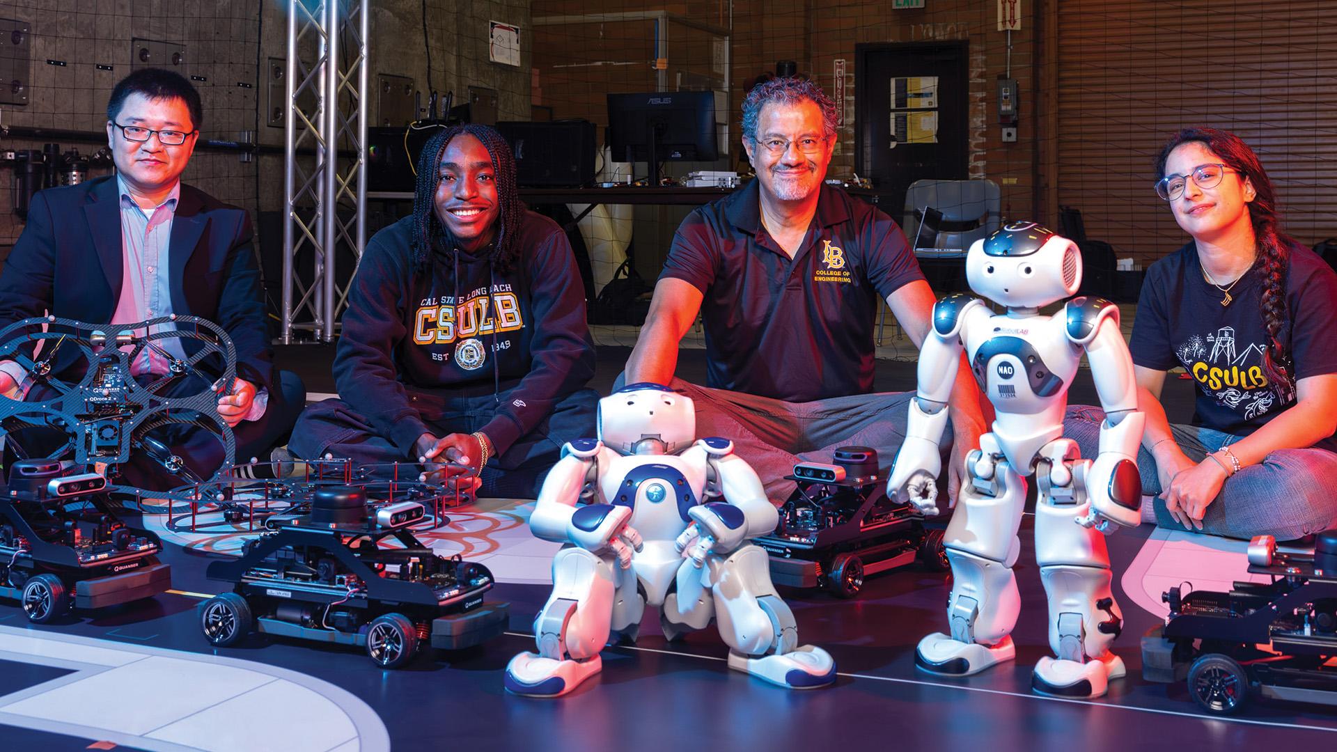 Five people sit on the floor in a robotics lab surrounded by small vehicles, drones and two humanoid robots.