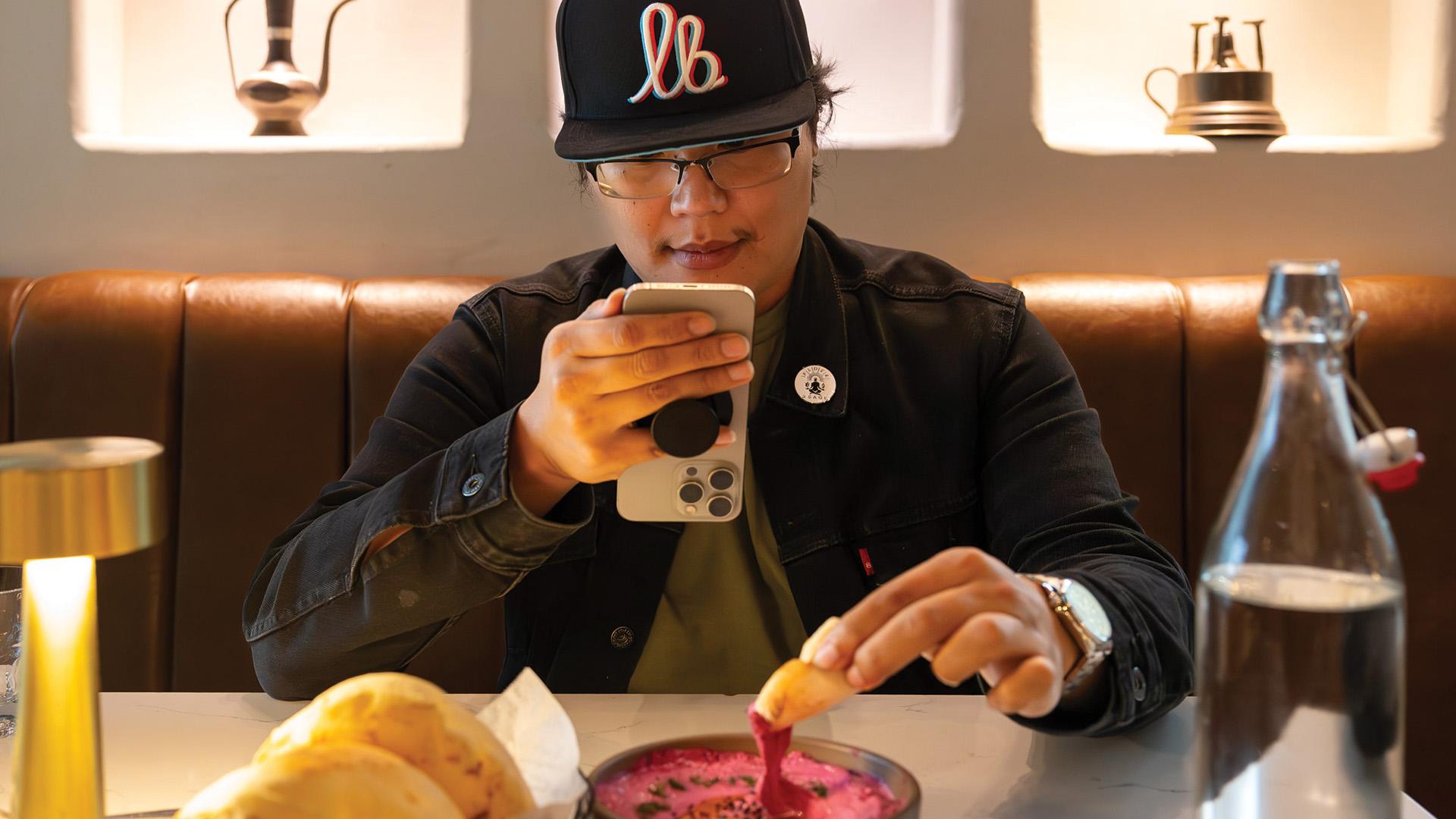 Person sits in a booth photographing a bowl while dipping bread into it.