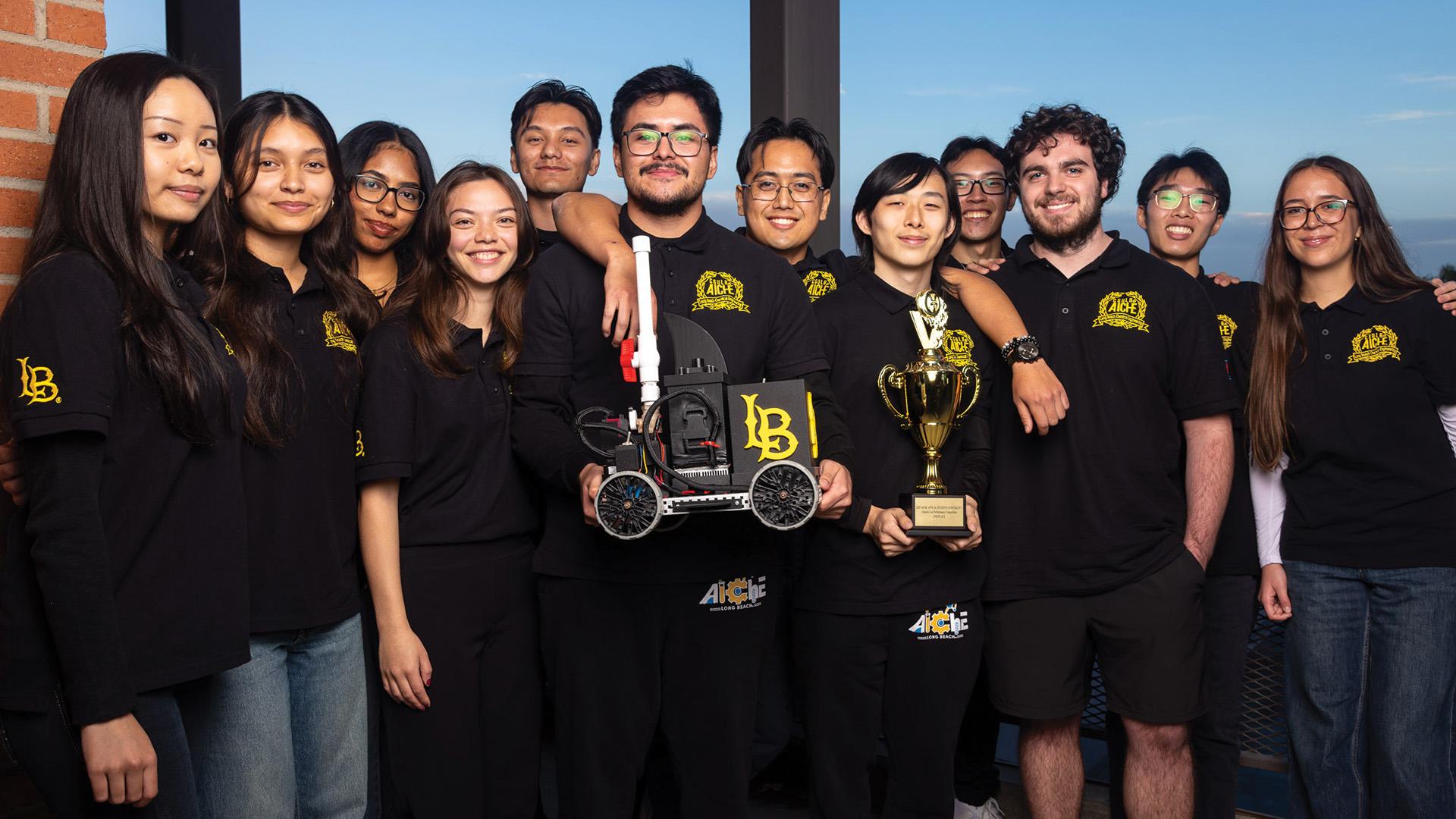 A group of CSULB students stands together as two hold a robotic car and a trophy.