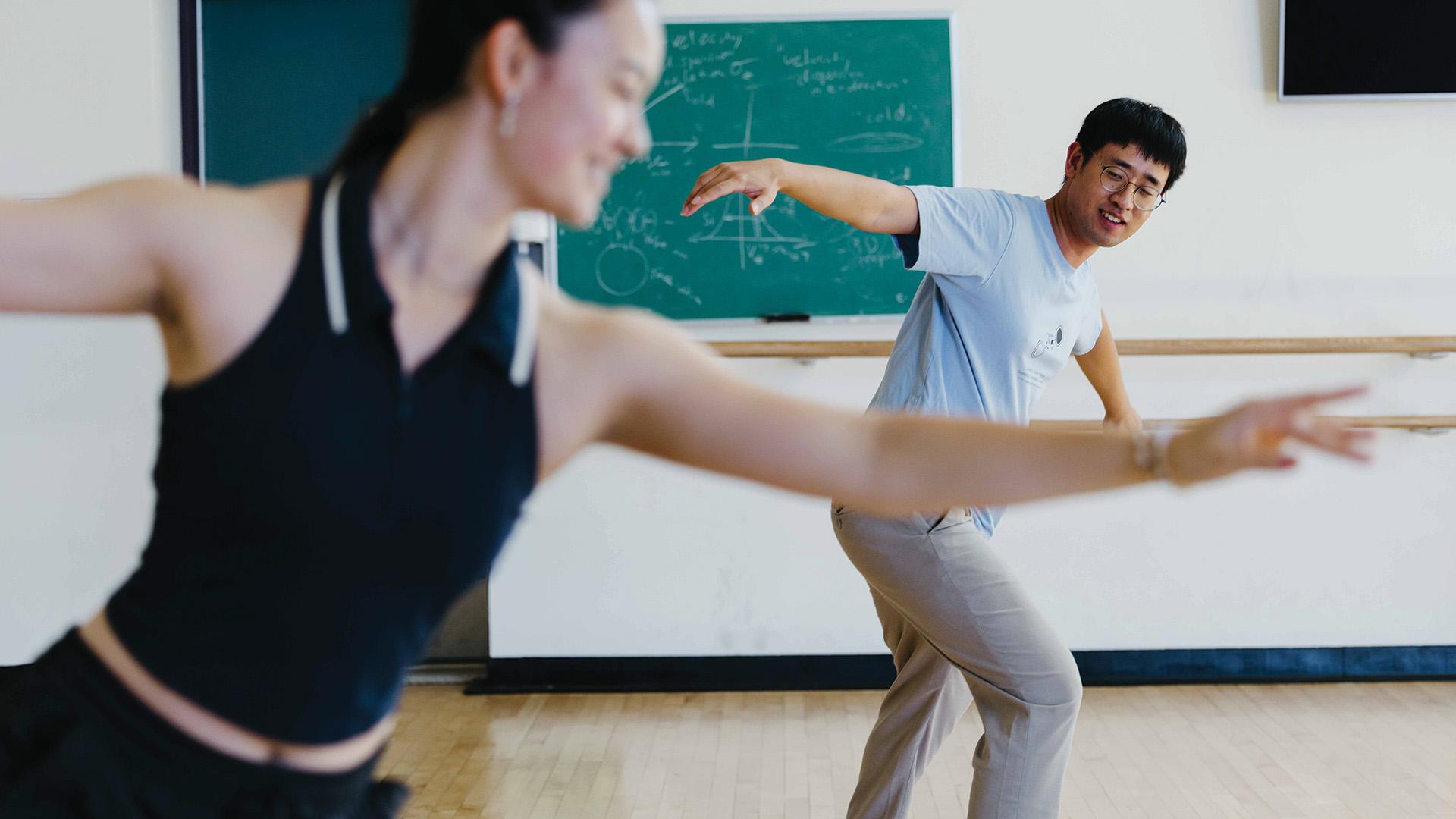 Two people dance in front of a chalkboard covered in physics equations