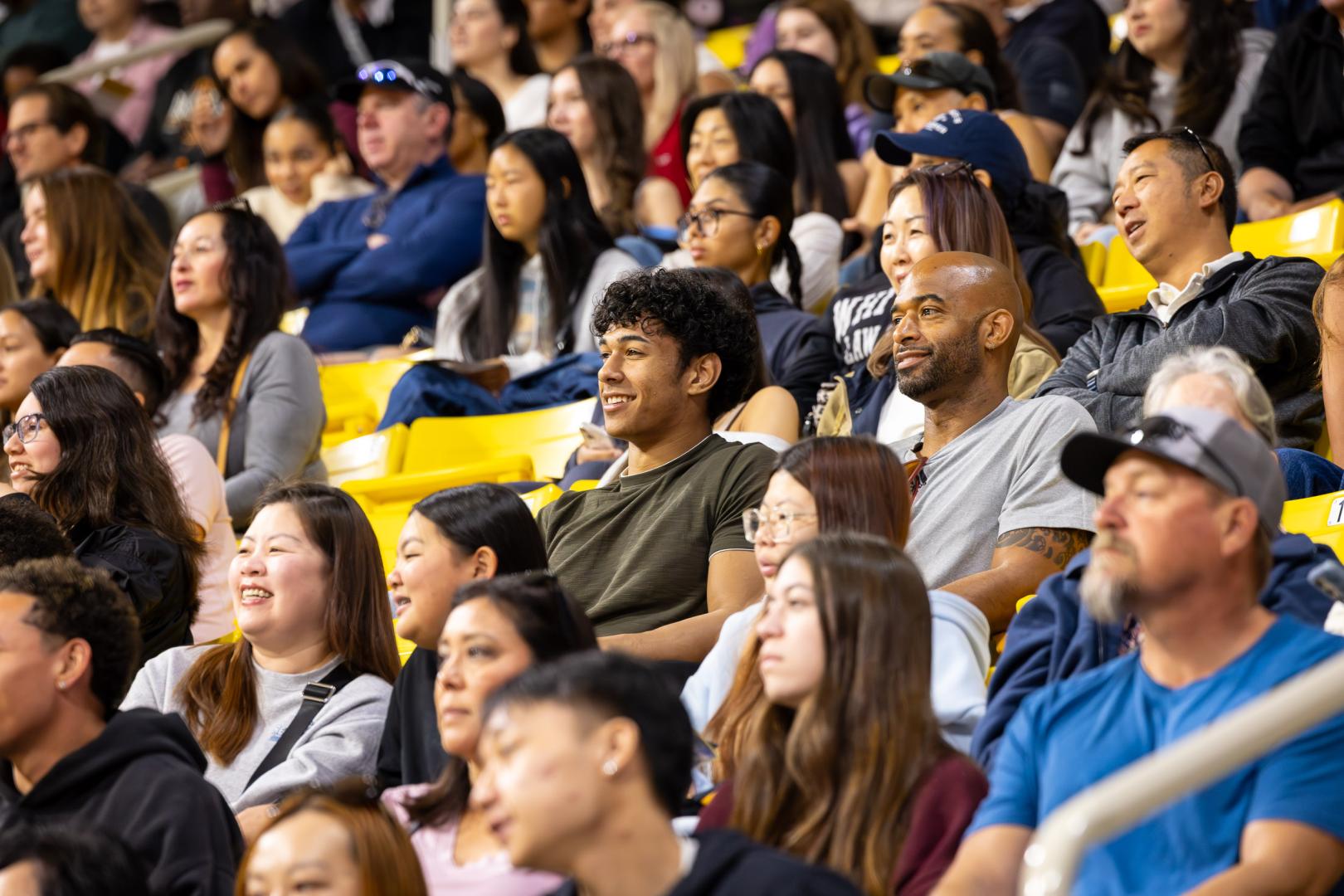 A large, diverse audience sits in bright yellow bleachers inside an indoor arena. Several spectators are smiling and watching an event, while others are talking or looking toward the floor. In the center, a young man in a dark green shirt smiles beside a man in a gray T-shirt. The foreground and background are softly blurred, emphasizing the central spectators.