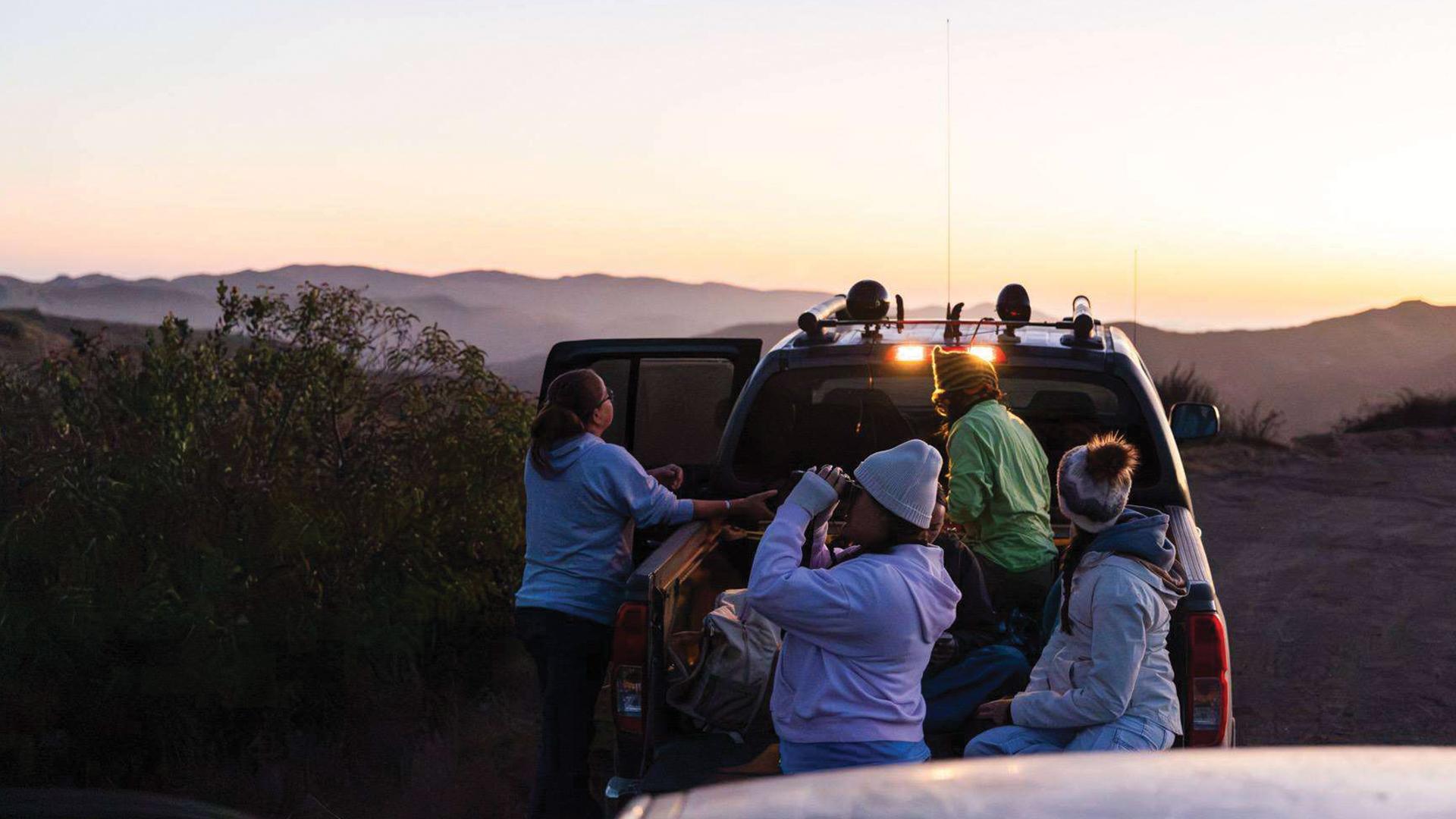 Students wearing hats and sweatshirts gather at dusk in a hilly area