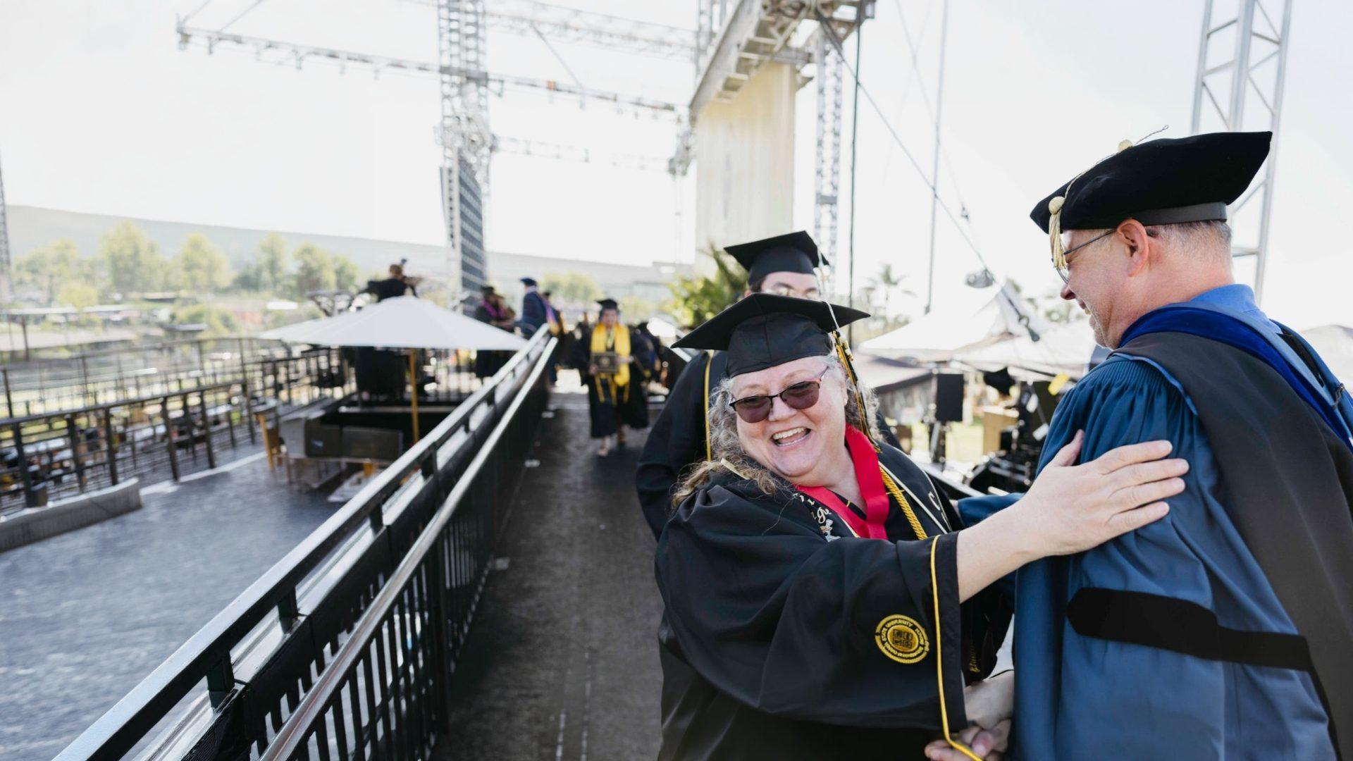 A smiling Cal State Long Beach graduate in cap and gown greets a faculty member in doctoral robes at commencement