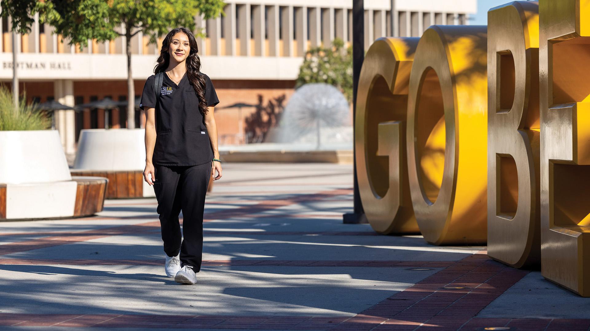 A person in nursing scrubs walks past giant “Go Beach” letters on campus.