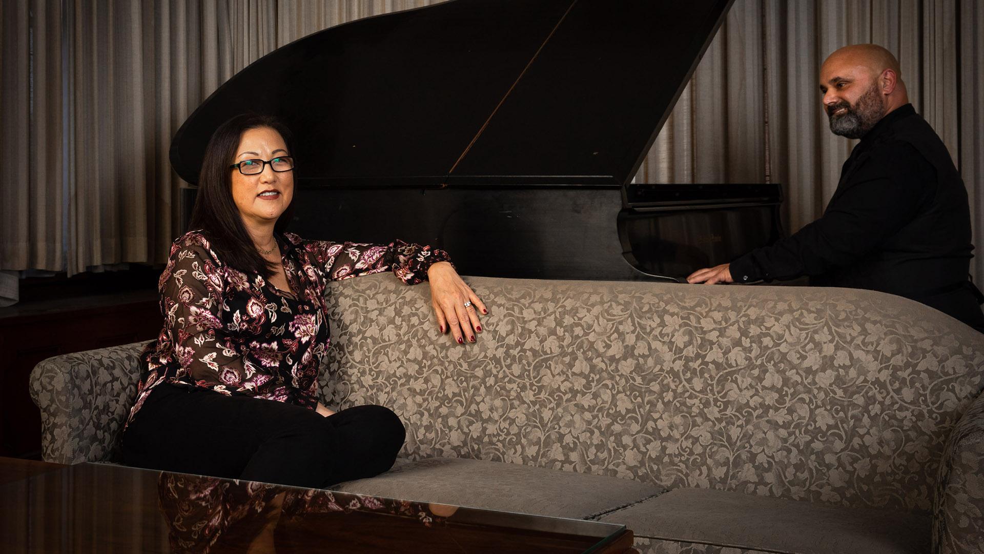 Portrait of woman sitting on couch, with man playing piano behind her. 