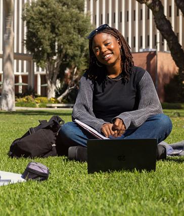 A smiling student with laptop in front of her sitting on a campus lawn