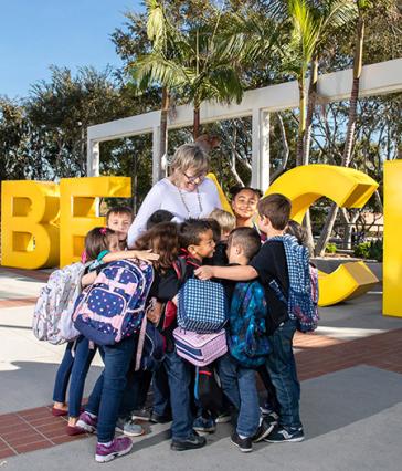Long Beach State University President Jane Close Conoley stands among a group of children with backpacks who are hugging her in front of large yellow "GO BEACH" letters on campus.