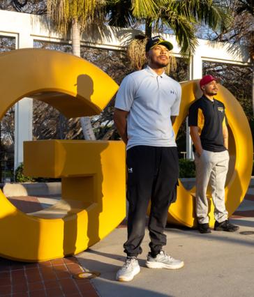 Alumni Markel and Martel Cooper stand in front of large yellow "GO BEACH" letters at Long Beach State 