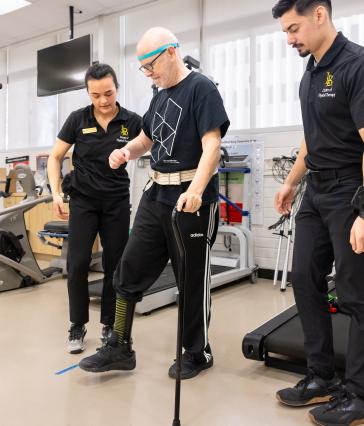 Two students helps a Neuro Lab client as he uses a cane to walk