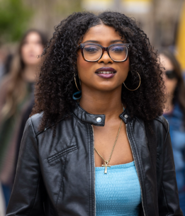 A person with curly hair, glasses and a leather jacket looks ahead while walking outdoors