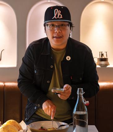 Person stands at a restaurant table holding a phone while preparing to eat from a bowl.