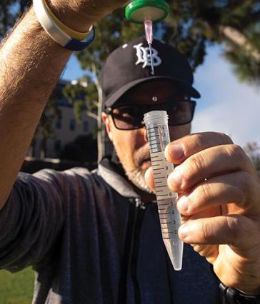 A person holds a syringe over a clear tube, releasing drops of liquid during outdoor sample collection.