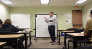 Harman teaching A man stands in front of a white board, teaching a class, with a marker in his hand.