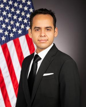 Mentor Victor Luquin wearing a suit and standing in front of an American flag