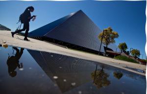A student carrying a waterbottle and looking at their phone walks in front of Walter Pyramid. In the foreground, there is a sizeable puddle.