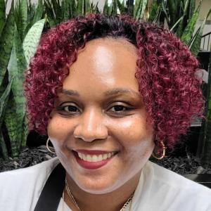 Nakia Thierry '14 Person with short curly hair smiles in front of tall leafy plants.