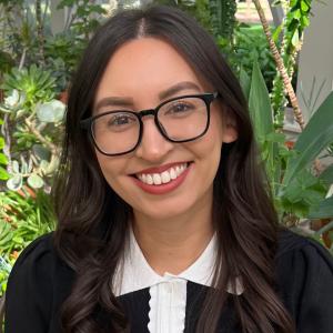 Desirae Moncayo '17 Person with glasses and long hair smiles in a plant-filled room.