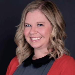 Melissa Dillon '07 Person with curled hair and cardigan smiles in front of dark background.