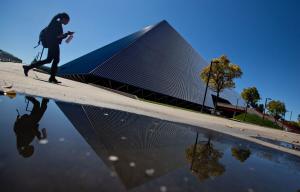 A student carrying a waterbottle and looking at their phone walks in front of Walter Pyramid. In the foreground, there is a sizeable puddle.