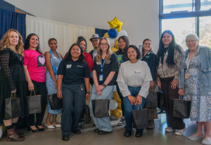 A line of various community partners are standing in a row in front of a backdrop and balloons. There are 12 of them that are holding black gift bags.