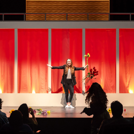 A Theatre Arts performer A performer stands onstage with arms outstretched, holding a yellow flower, while a silhouette in the audience throws flowers toward the stage.