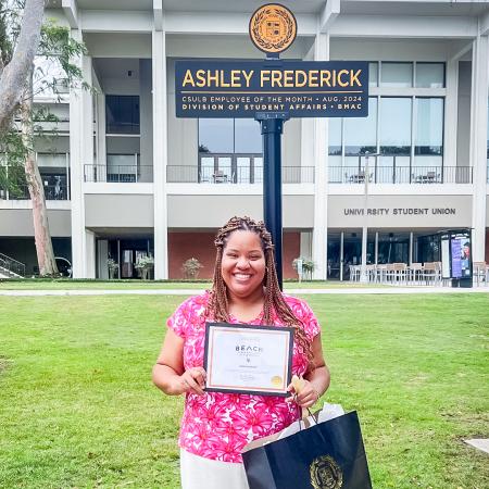 Ashley Frederick holds an award while standing under a sign that reads "Ashley Frederick, Employee of the Month."