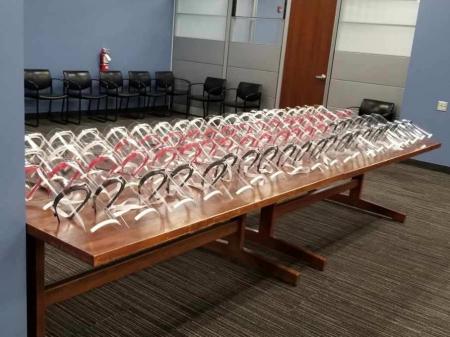 A large wooden table filled with rows of 3D-printed clear plastic face shields, arranged neatly in a conference room. A large wooden table filled with rows of 3D-printed clear plastic face shields, arranged neatly in a conference room.