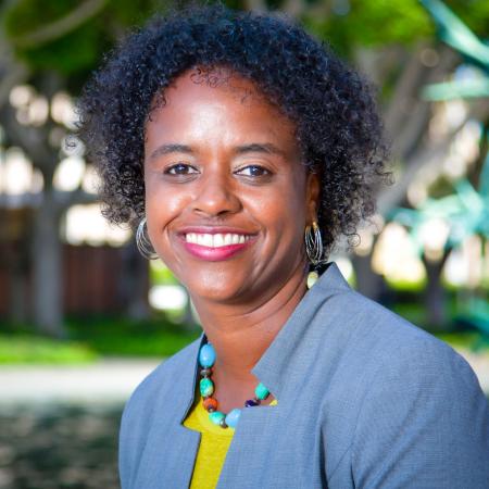 Rashida Crutchfield A person smiles at the camera, wearing a gray blazer and colorful necklace, standing outdoors.