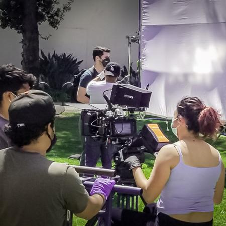 Several people on a film set, one of whom operates a camera, all wearing masks and working with film equipment. Several people on a film set, one of whom operates a camera, all wearing masks and working with film equipment.