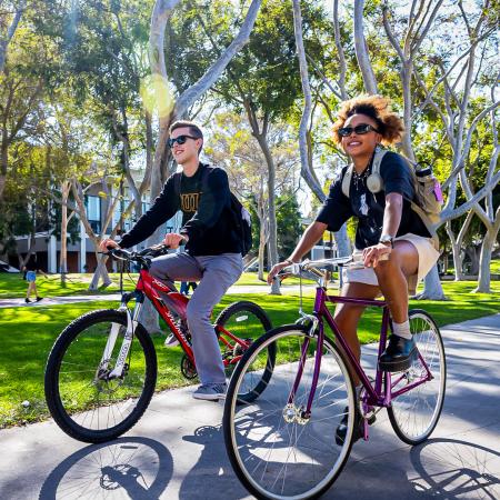 Two students ride bikes along a campus path surrounded by trees. Two students ride bikes along a campus path surrounded by trees.