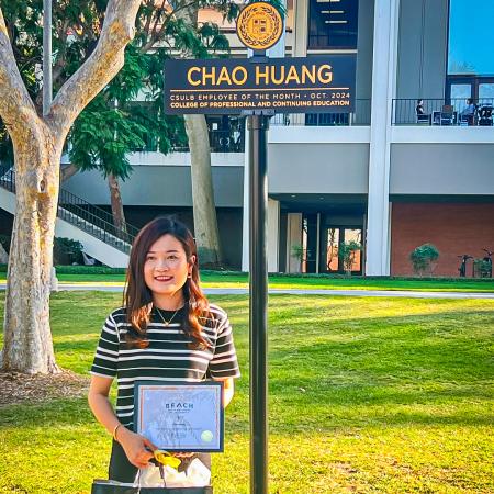 October Employee of the month Chao Huang Chao Huang stands holding a certificate and bag under a sign bearing her name.