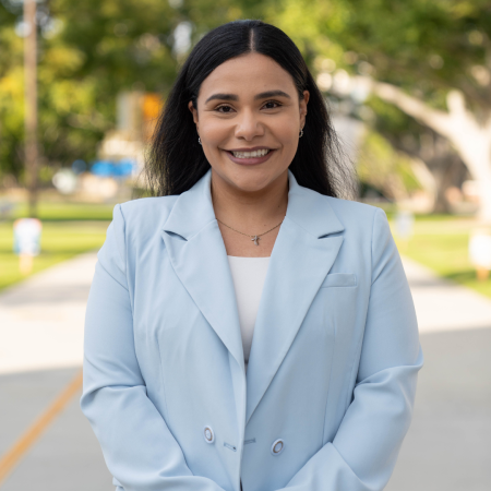 Celia Mejja, of the Women's and Gender Equity Center Celia Mejia smiles outdoors in a light blue blazer, with trees and a path in the background.