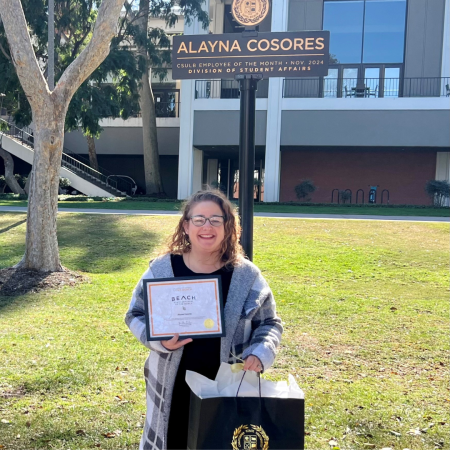 Alayna Cosores, Employee of the Month in November 2024 Alayna Cosores stands smiling outdoors holding a framed certificate and gift bag beneath a sign with her name on it.