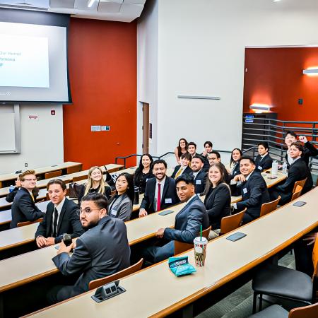 A group of business students in formal attire sit in a lecture hall, smiling at the camera. A group of business students in formal attire sit in a lecture hall, smiling at the camera.