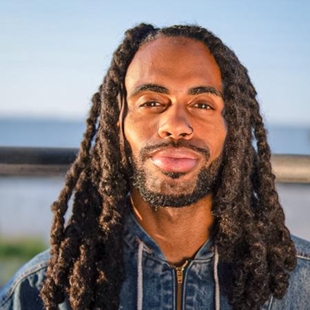 Assistant Professor Danzel Thompson-Stout smiles softly while wearing a denim jacket against a blurred outdoor background. Assistant Professor Danzel Thompson-Stout smiles softly while wearing a denim jacket against a blurred outdoor background.